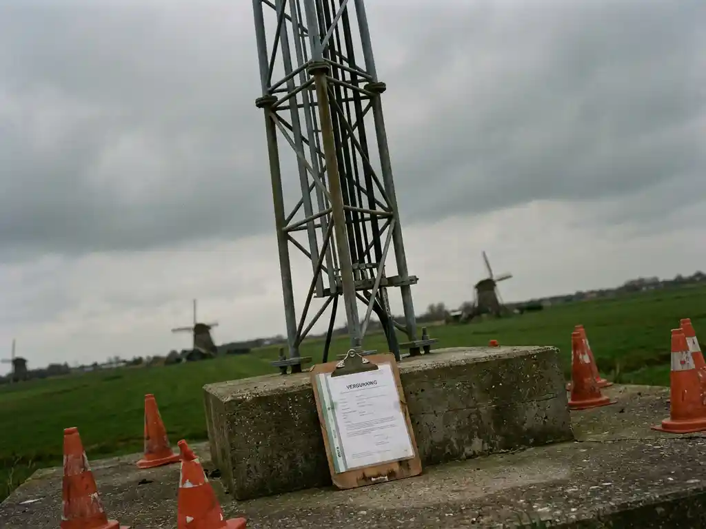 Mobiele zendmast met veiligheidskegels en vergunningsdocumenten aan de basis, Nederlandse landschap met windmolens op achtergrond